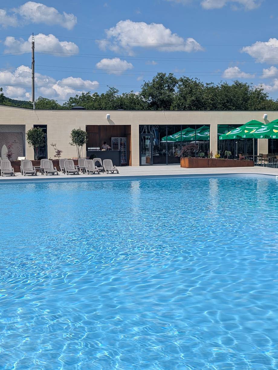 Modern poolside terrace with loungers, green umbrellas, and service building under blue sky