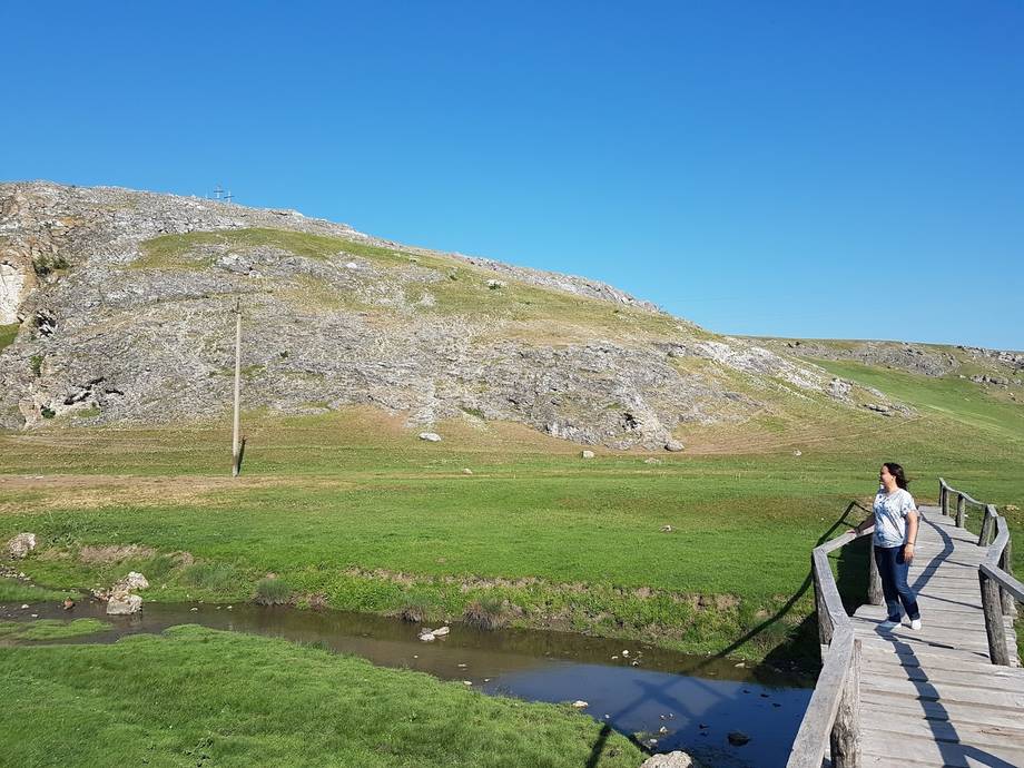 Wooden boardwalk crossing a stream through rolling limestone hills and green pasture
