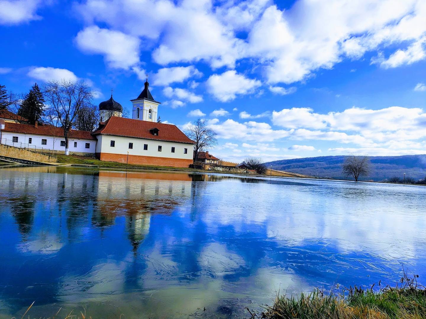 Waterfront monastery with church steeple reflected in calm blue water, mountains beyond