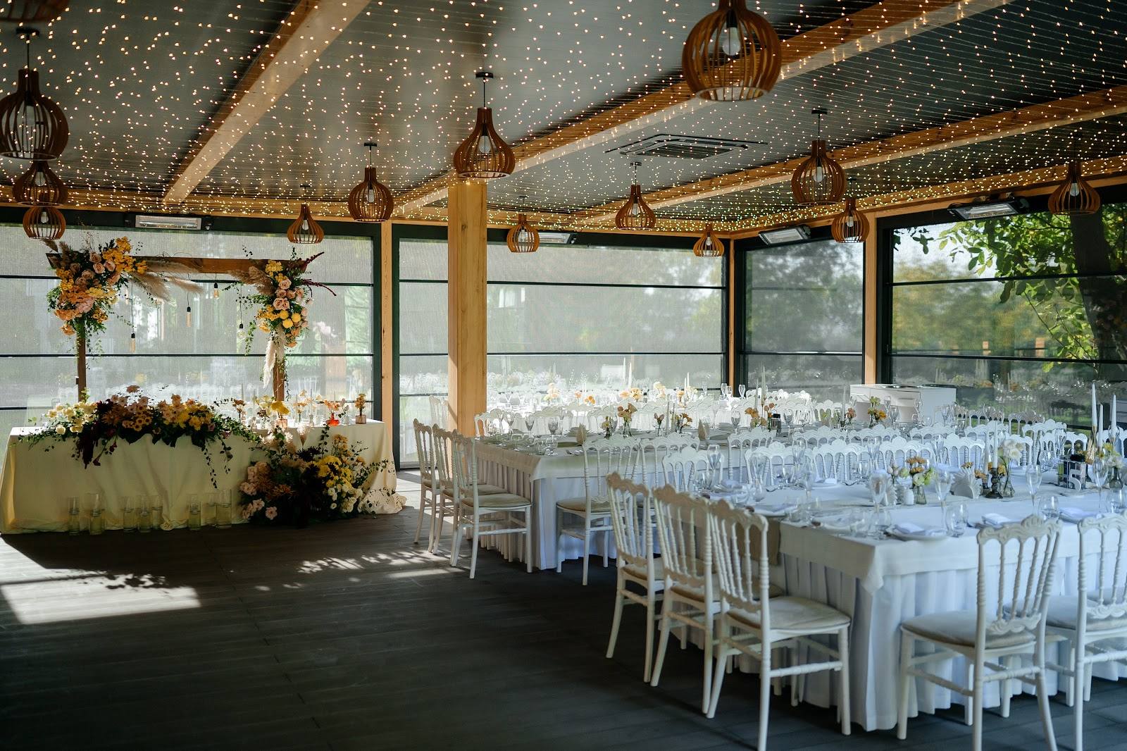 Elegant event venue with wooden beams, white chiavari chairs, and decorated floral arrangements ready for reception