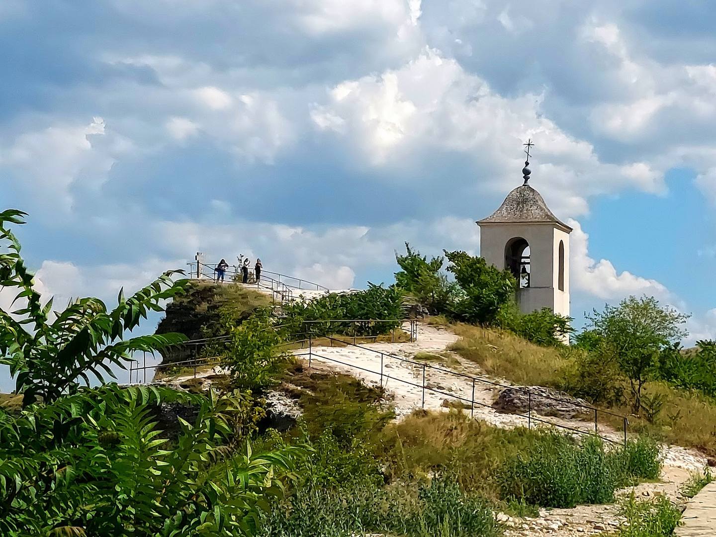 Small chapel with bell tower atop a grassy limestone hillside under dramatic clouds