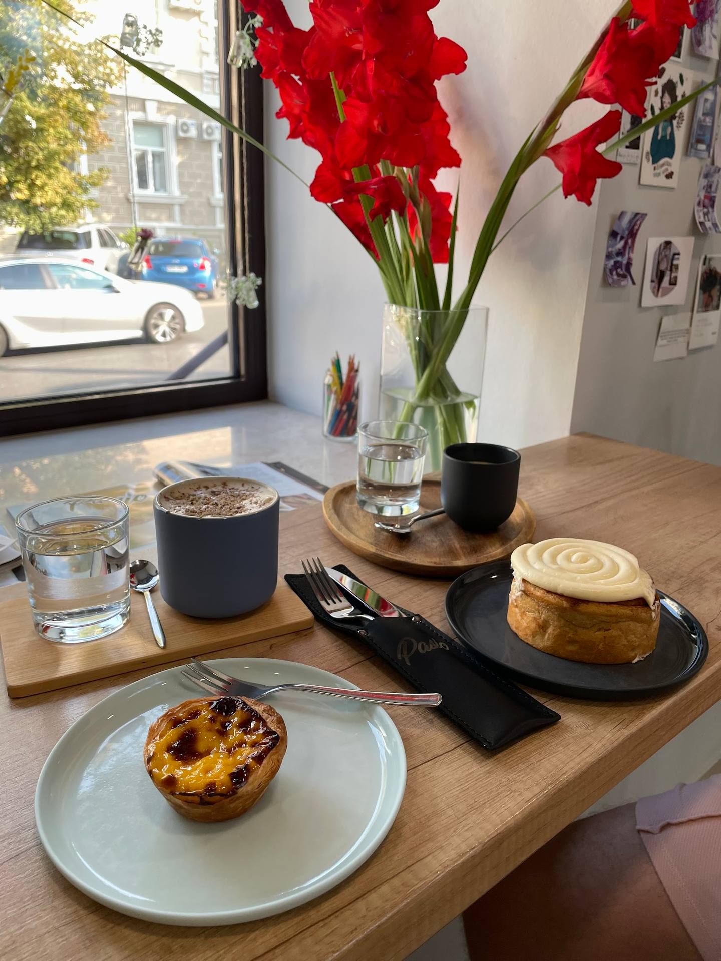 Pastries and beverages on wooden table with red gladiolus arrangement and street view