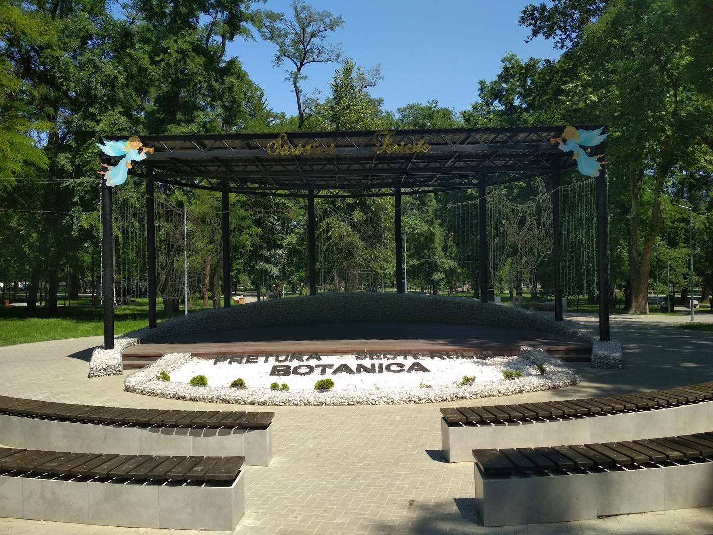 Open-air event pavilion with metal pergola structure and signage in landscaped park setting
