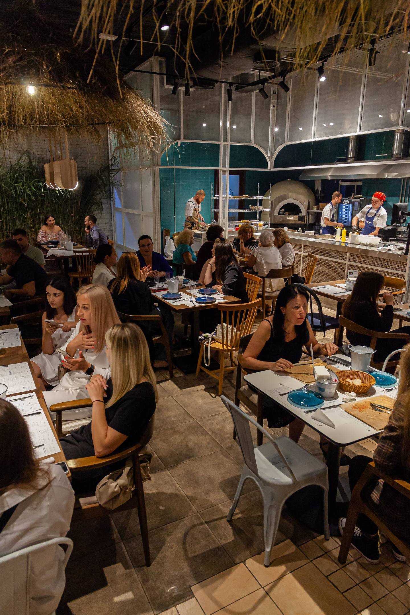 Modern restaurant interior with open kitchen, dining area with guests enjoying meals, and thatched ceiling details