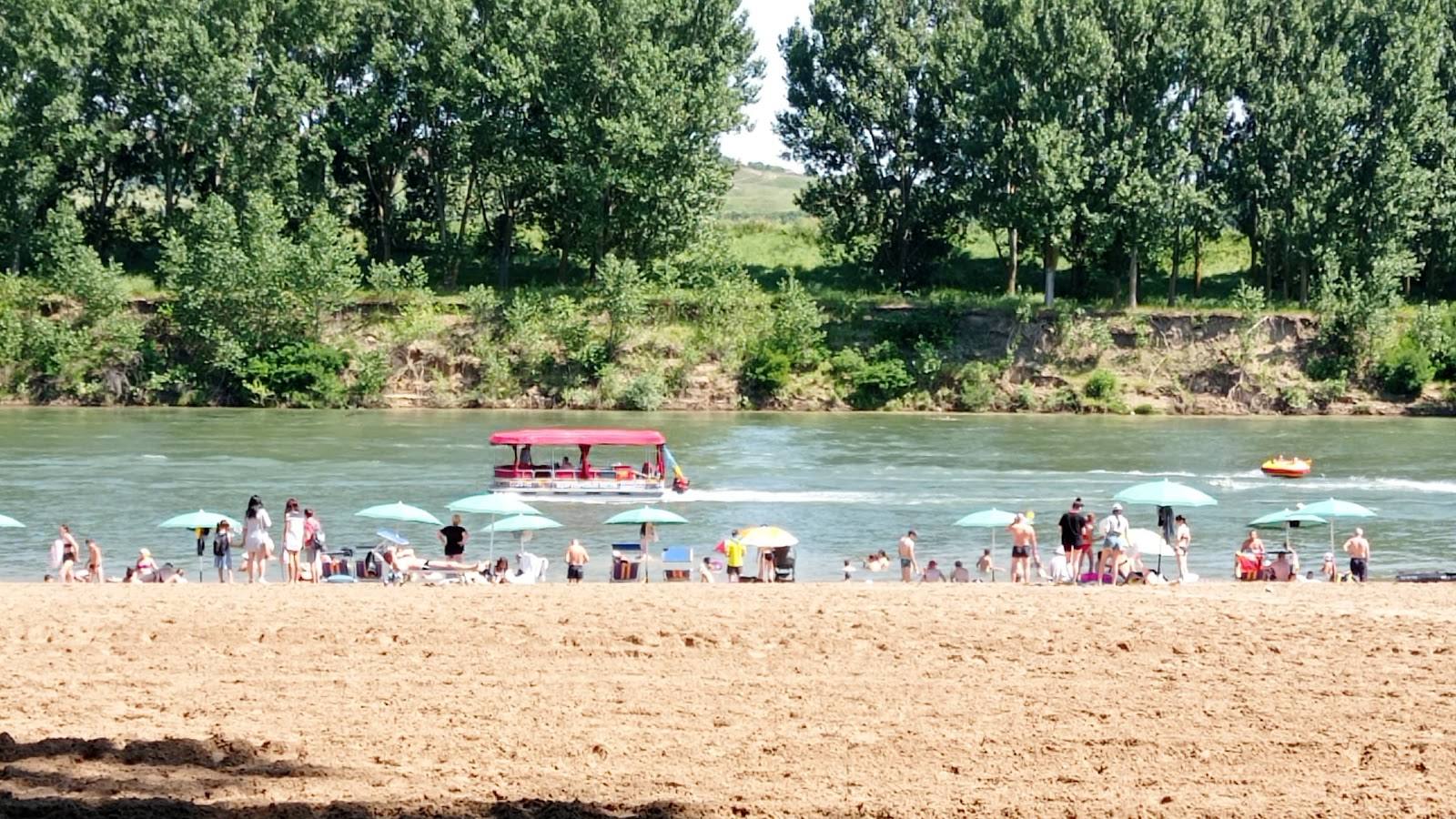 Sandy riverside beach with visitors, floating bar, and green valley backdrop