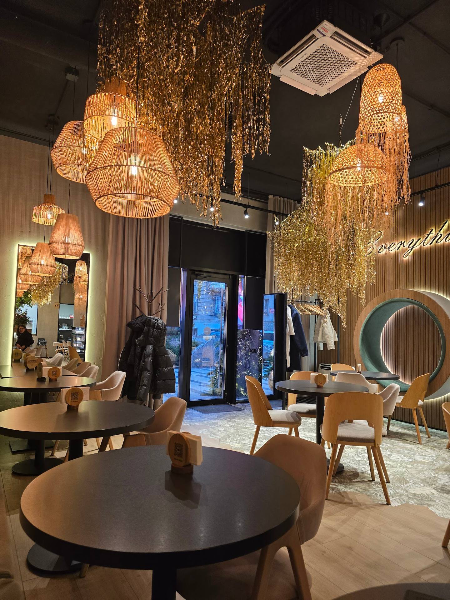 Modern restaurant interior with sculptural pendant lighting, round dining tables, and floor-to-ceiling storefront at dusk.