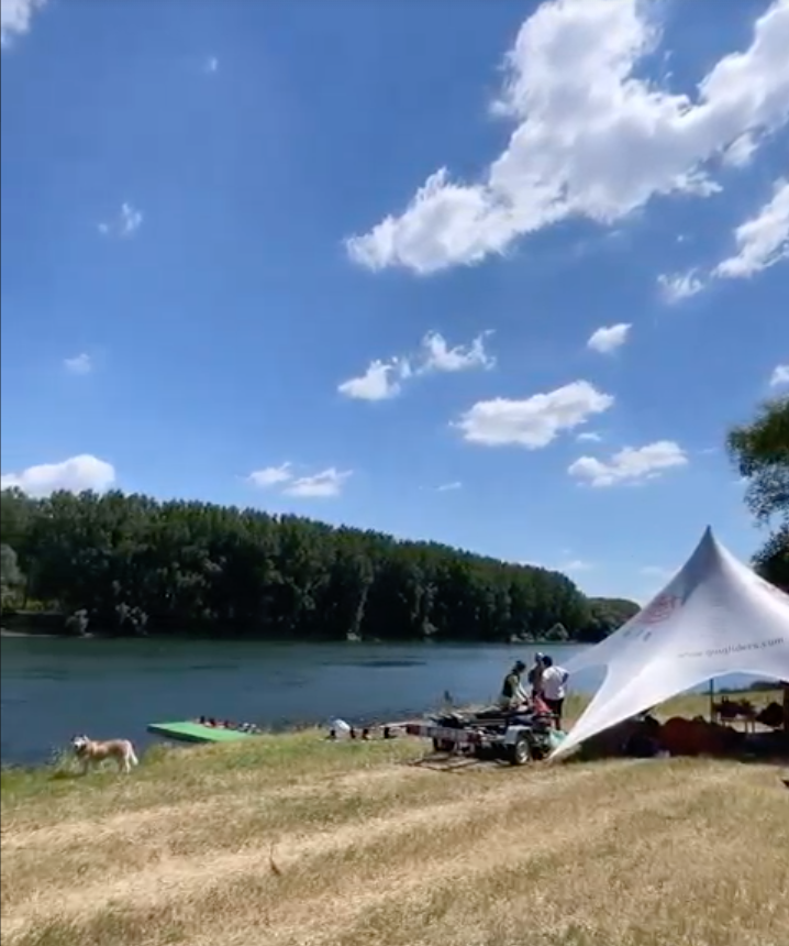 Riverside event venue with white tent setup, forest backdrop, and sandy waterfront grounds
