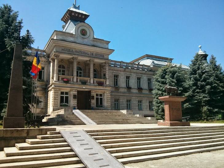 Neoclassical government building with clock tower and ceremonial entrance steps, framed by monument and flagpole
