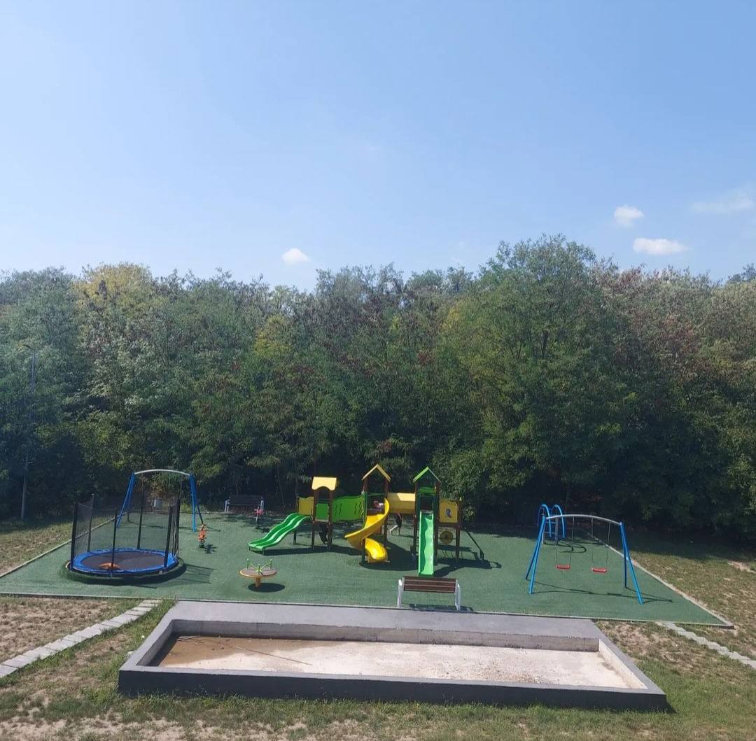 Outdoor playground with slide, climbing equipment, sandbox and seating area surrounded by trees