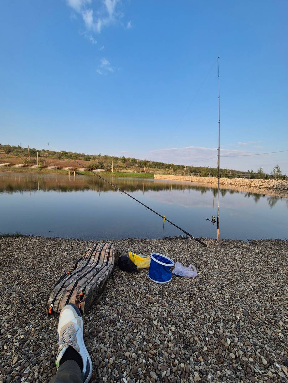 Fishing setup with rod, tackle, and calm reservoir under clear sky