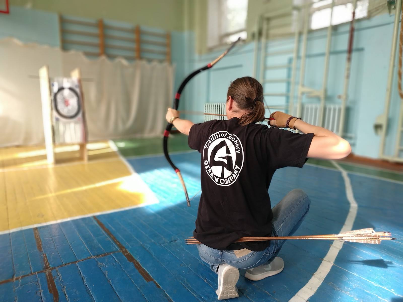 Archer practicing archery indoors on blue gym floor with target visible in background