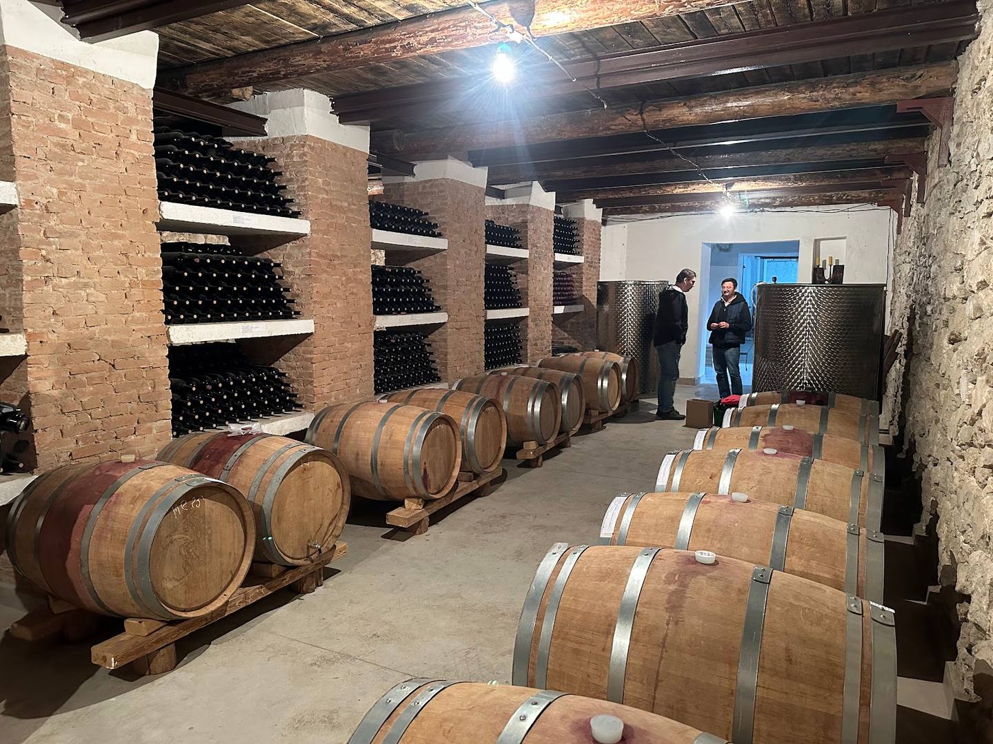 Wine cellar with wooden barrels and bottle storage shelves along brick walls