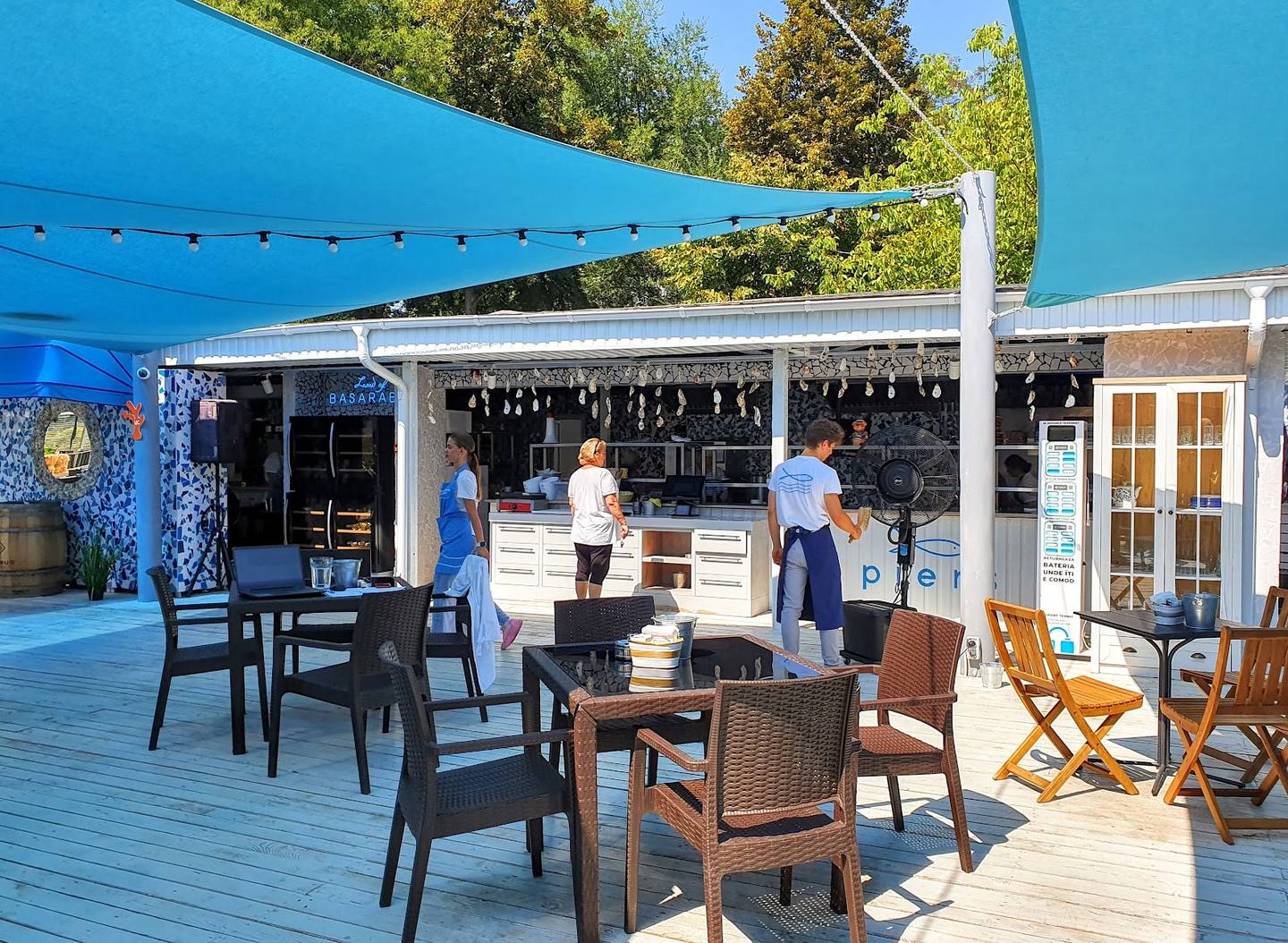 Outdoor terrace with blue shade sail, bar counter, and mixed seating under clear sky