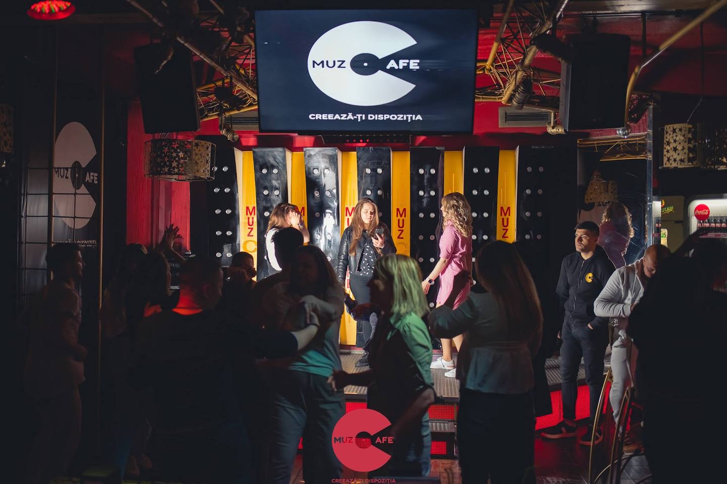 Dimly lit arcade/game bar interior with illuminated cabinet games and visitors enjoying the space