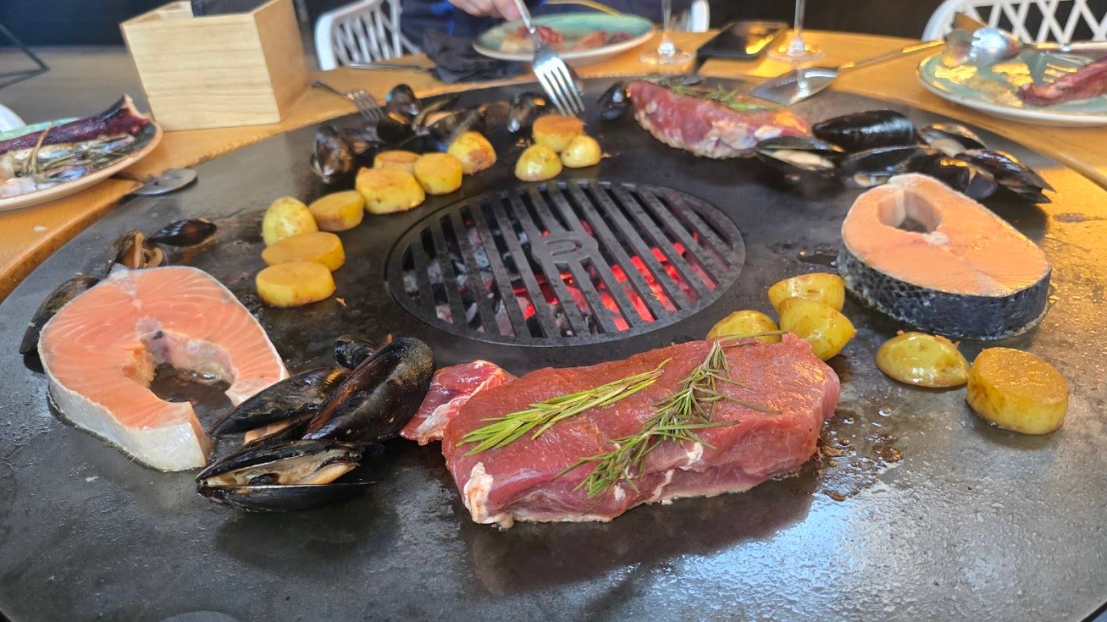 Table-top grilling setup with raw meat, seafood, and vegetables on a stone surface