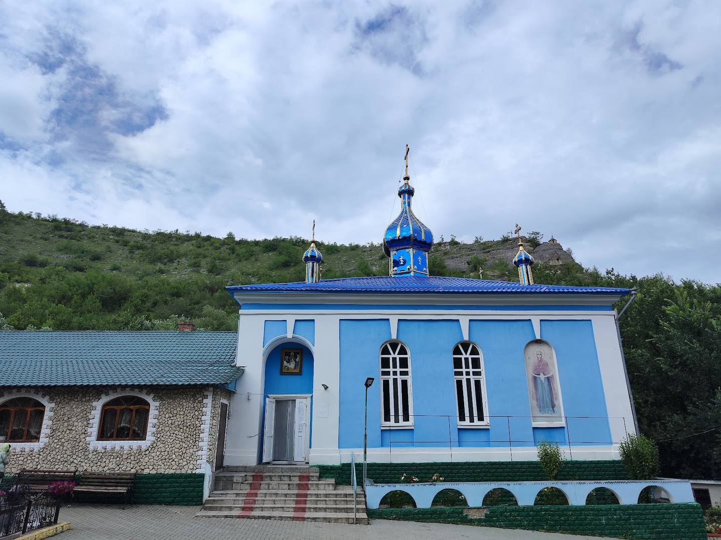 Bright blue monastery chapel with ornate domes and cross, nestled in green hillside landscape