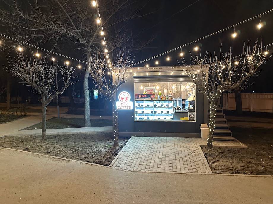 Illuminated kiosk storefront at night with festive string lights and landscaping