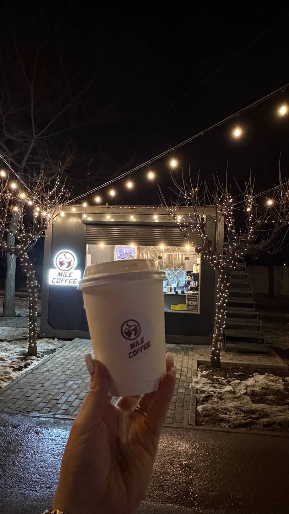 Branded coffee cup held up in front of nighttime storefront with festive lighting