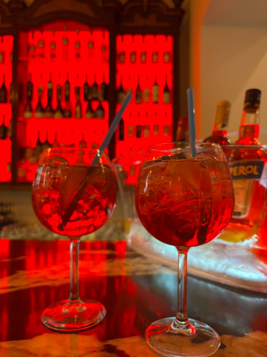 Two vibrant red cocktails with ice and straws at a bar with illuminated backlit bottle display