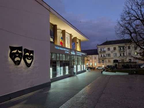 Theater storefront with theatrical masks signage, lit evening facade, urban square setting